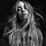 Sydney Sweeney, with long, loose hair squats barefoot in a wide-legged pose while wearing a denim jacket and flared jeans. She looks directly at the camera with a neutral expression. The background is a faded black-and-white photo of women and children.