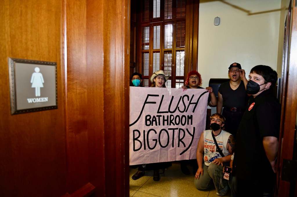 Several activists sit and stand inside a women’s restroom at the Texas Capitol, holding a large pink banner with black letters that read “Flush Bathroom Bigotry.” Some wear masks, and one person wears a cowboy hat.