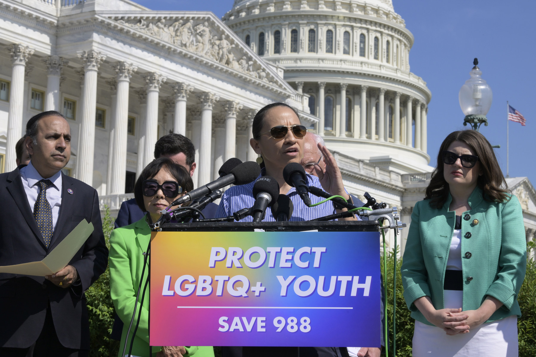 Politicians stand in front of a podium that says "protect LGBTQ youth save 988" with the Capitol building in the background.