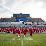 A women’s tackle football team, the Washington Prodigy, walks hand in hand across the field in red uniforms, with rows of teammates standing behind them and empty stadium bleachers in the background.