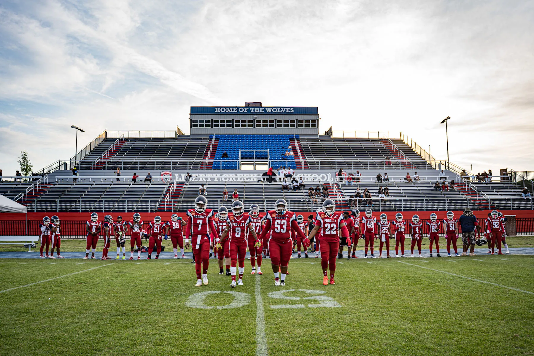A women’s tackle football team, the Washington Prodigy, walks hand in hand across the field in red uniforms, with rows of teammates standing behind them and empty stadium bleachers in the background.