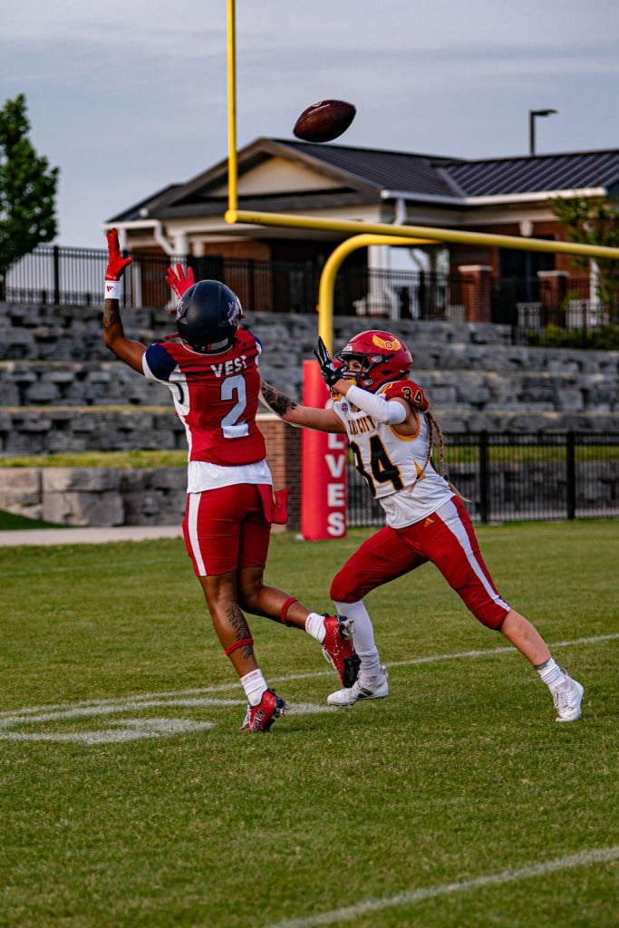 A Washington Prodigy receiver in a red and navy uniform reaches for a pass near the end zone while being closely defended by an opposing player in a white and red uniform, with the goalpost visible behind them.