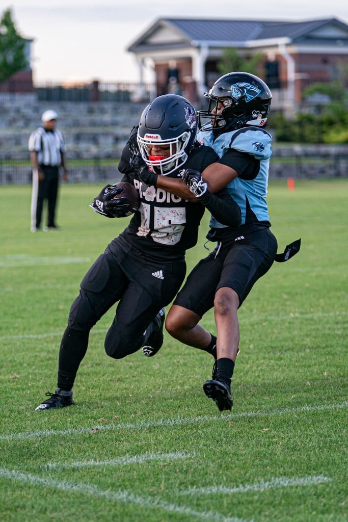 A Washington Prodigy player in a black uniform carries the football while being wrapped up and tackled by an opponent in a light blue and black uniform during a game.