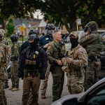 A group of armed law enforcement officers wearing tactical gear and face coverings stand together on a residential street lined with parked cars. Some wear camouflage uniforms and helmets, others wear dark uniforms with protective vests.