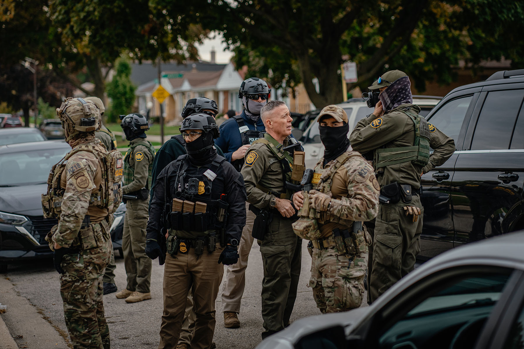 A group of armed law enforcement officers wearing tactical gear and face coverings stand together on a residential street lined with parked cars. Some wear camouflage uniforms and helmets, others wear dark uniforms with protective vests.