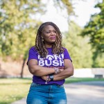 Cori Bush stands with her arms crossed in a park, wearing a purple campaign T-shirt and looking off to the side.