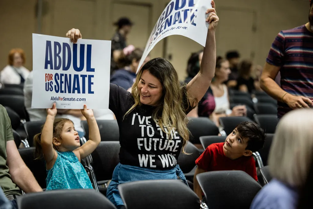 A woman and two children hold Abdul El-Sayed campaign signs inside a rally venue; the woman raises one sign overhead while smiling.