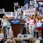Senate candidate Abdul El-Sayed smiles at a podium as supporters behind him raise campaign signs.