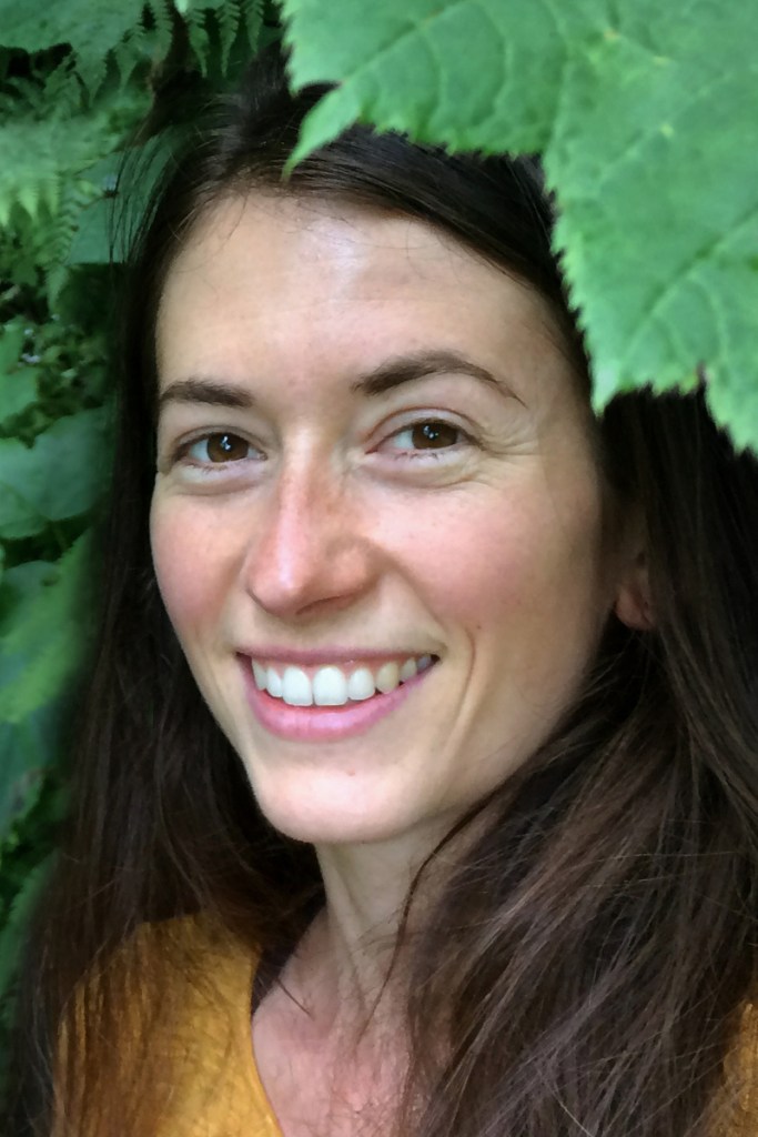 A woman smiles in a closeup portrait of her face surrounded by green leaves.