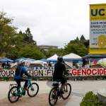 Two cyclists ride by a protest encampment with signage showing support for Palestine and a UCI sign on a light pole nearby.