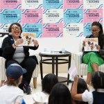 Two women laugh and read books to an audience of children while seated on the stage.