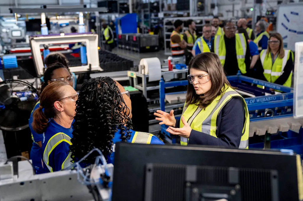 Haley Stevens, wearing safety glasses and a reflective vest, speaks with workers on a factory floor as other employees stand nearby.