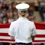 A woman in military uniform is seen from behind facing an audience and the US flag.