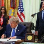 President Donald Trump signs a document with a black pen, with a woman holding her pregnant belly to the left of him and Robert F. Kennedy Jr. to the right.