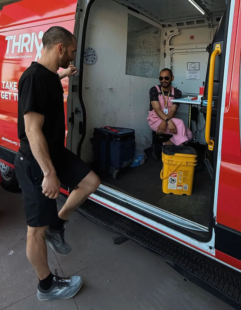 One outreach worker sits inside a red THRIVE mobile health van while another stands outside talking with him.