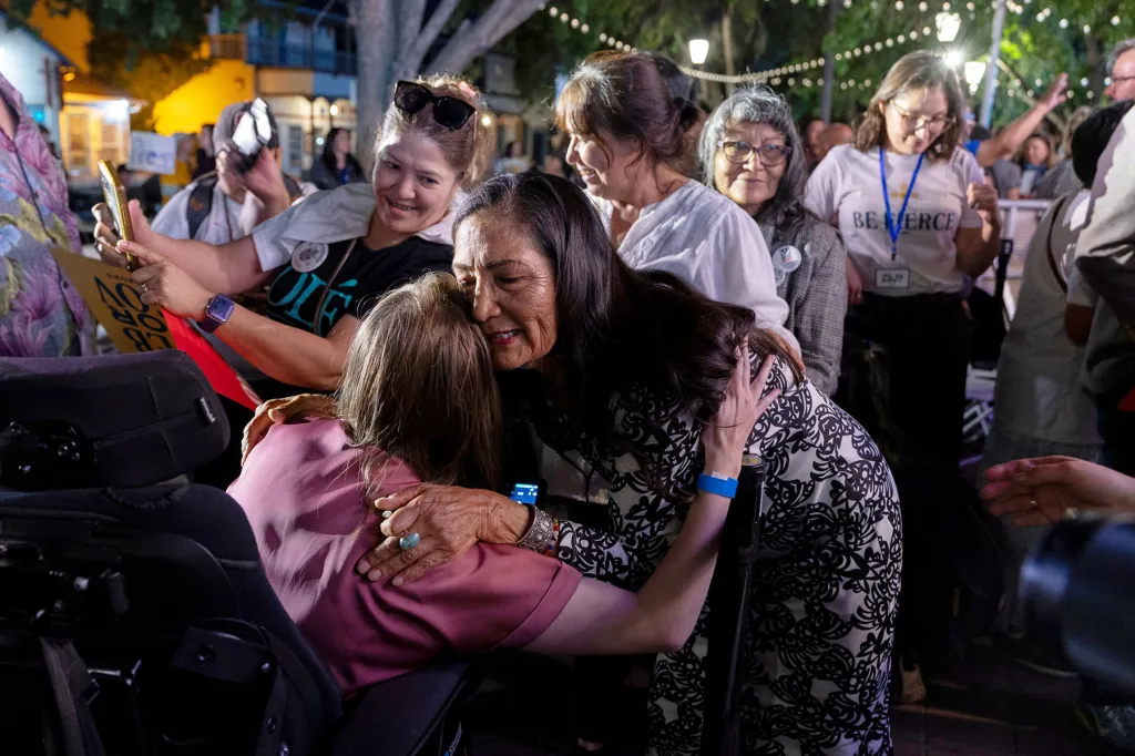 Deb Haaland embraces a supporter in a crowd at an outdoor evening event as other attendees gather around them.
