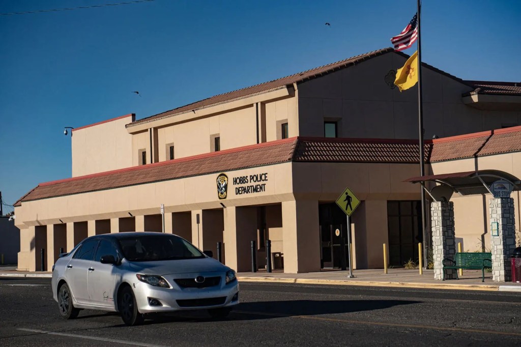 A car passes the Hobbs Police Department beneath American and New Mexico flags.