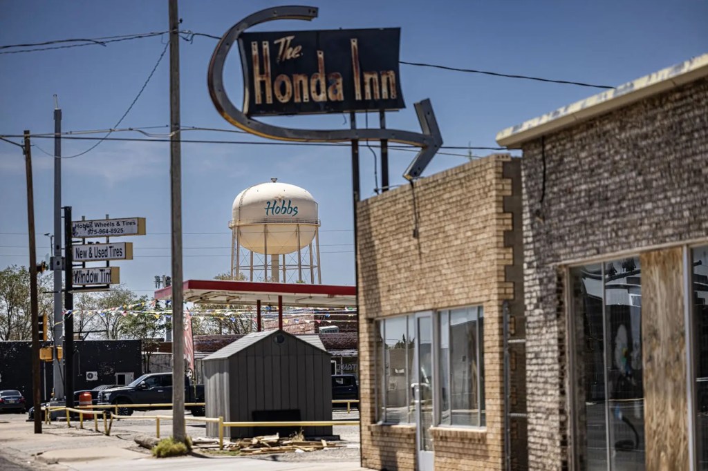 A vintage “The Honda Inn” sign rises above a street in Hobbs, New Mexico, with the city’s water tower and tire shop signs visible in the background.