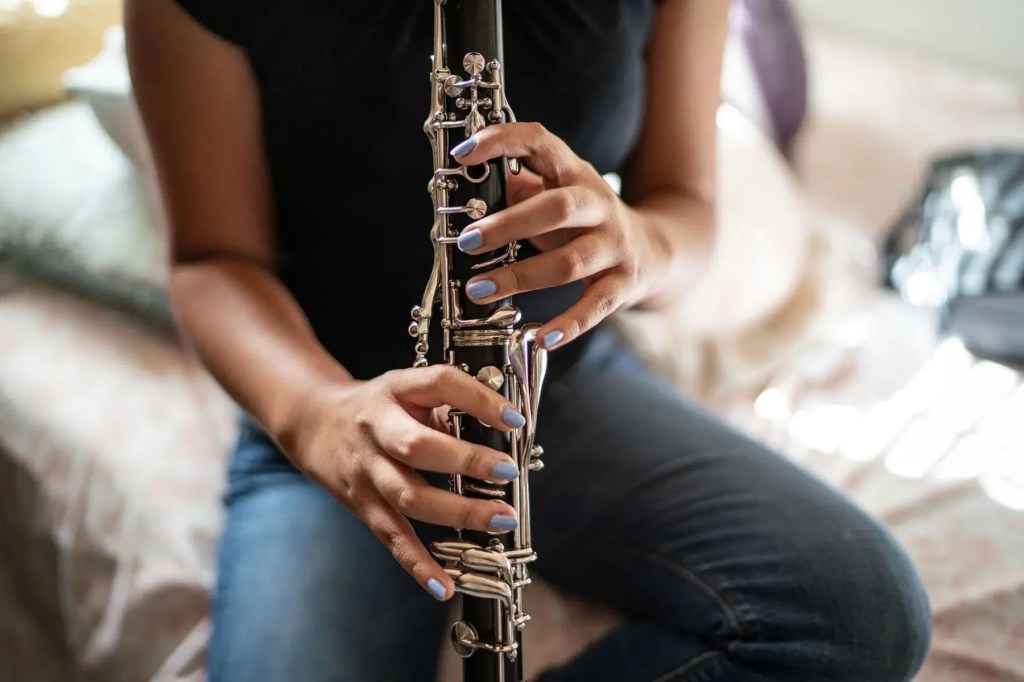 Close-up of a young girl’s hands, with light blue painted nails, holding a clarinet while seated on a bed.