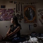 A young girl sits on a bed in a dark bedroom as narrow bands of sunlight from window blinds fall across her face and body.