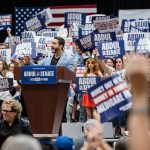 Senate candidate Abdul El-Sayed smiles at a podium as supporters behind him raise campaign signs.