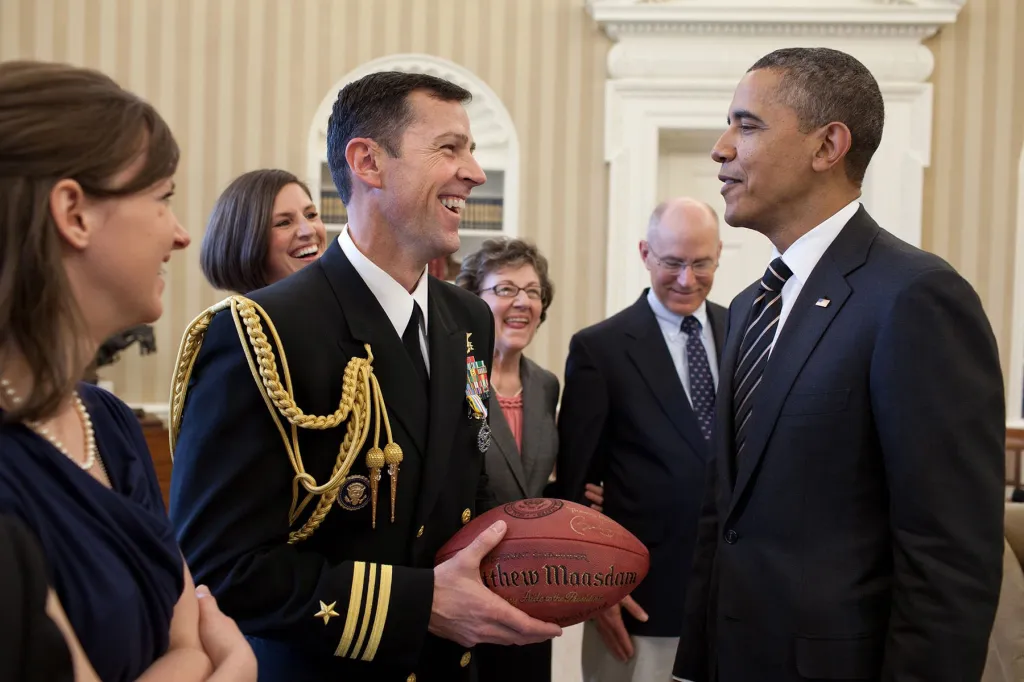 Matt Maasdam, wearing a Navy dress uniform and holding a football, smiles at President Barack Obama as several people look on in the Oval Office.