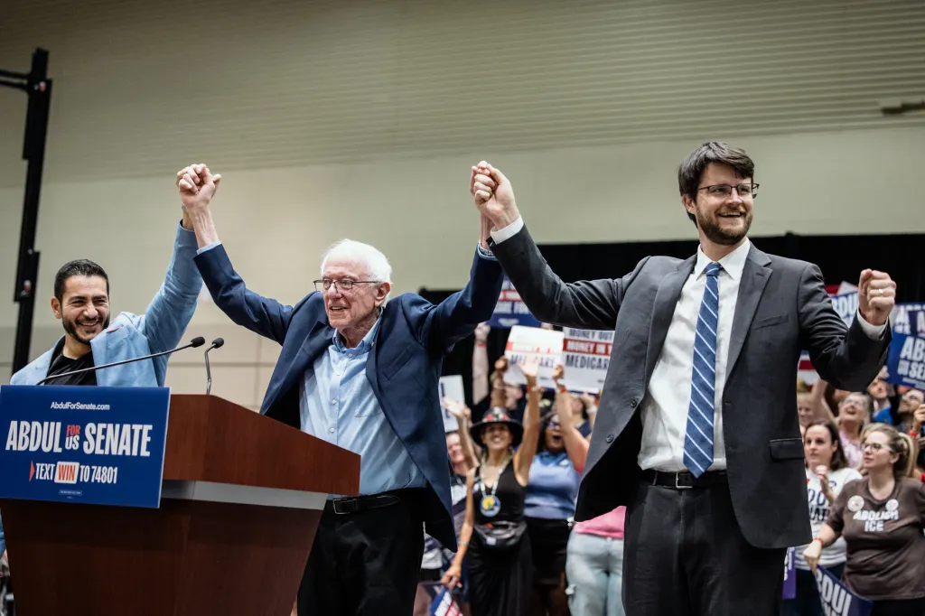 Abdul El-Sayed, Bernie Sanders and Will Lawrence stand onstage with their joined hands raised as supporters cheer behind them.