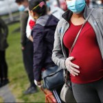 A pregnant woman wearing a mask and gloves stand outside among others in masks.