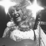 Parton ringed by lights on stage singing and playing guitar.