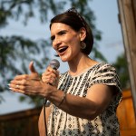 Peggy Flanagan speaks into a microphone at an outdoor event, gesturing with one hand as she addresses the crowd.