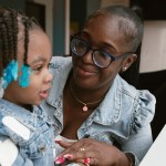 A woman wearing glasses and a denim jacket smiles down at a young girl wih braided hair adorned with blue bow barrettes, alson in a matching denim jacket, as the two share a close moment together indoors.