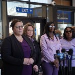 A group of women, several wearing purple, including Keshia Golden stand behind microphones during a press conference.