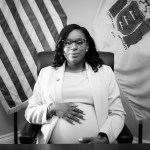 A portrait of McIver in an office surrounded by flags and holding her pregnant belly.