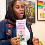 A person holds up a brochure about signs of a healthy relationship in a room with educational materials on the wall behind her.
