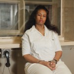 Kwaneta Harris in a white prison uniform sits behind a glass visitation partition with her hands cuffed in her lap. Two black telephone receivers hang on the wall beside her.