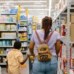 A woman and child walk down a store aisle stocked with household goods, including toilet paper and other everyday essentials.
