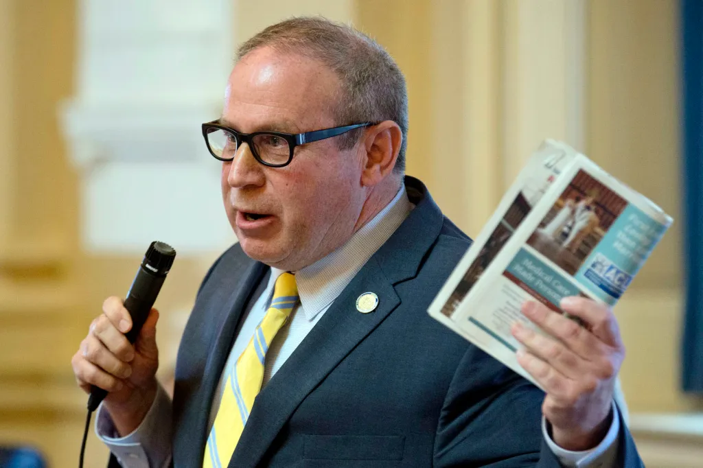 A man in glasses and a suit speaks into a microphone while holding up a printed document.