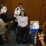 Lindsay Clancy is rolled into a courtroom in a wheelchair with her hands clasped in her lap as a uniformed officer stands behind her. Other court personnel and attorneys are visible around the room.