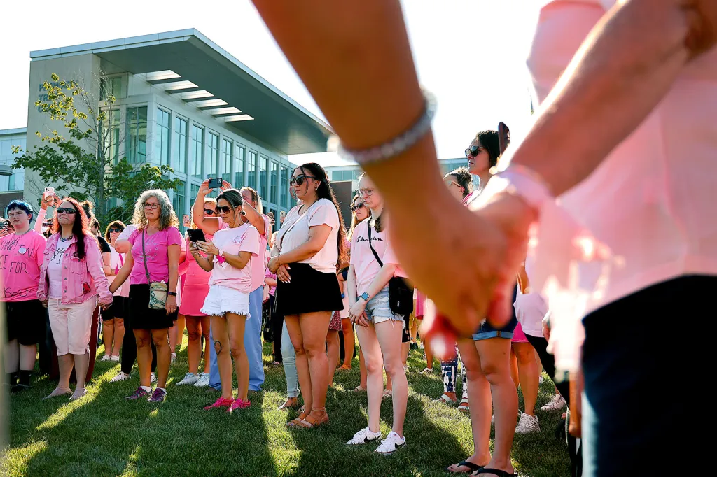 A crowd, many dressed in pink, gathers outside a courthouse in bright sunlight. Several people hold phones while two people in the foreground hold hands.