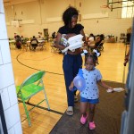 A woman carrying several takeout containers walks out of a gym with a young girl. People sit at tables in the background.