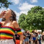 A person in a rainbow-striped off-the-shoulder top cups their hand around their mouth while shouting at a gathering. A crowd stands behind them beneath trees and a bright blue sky.