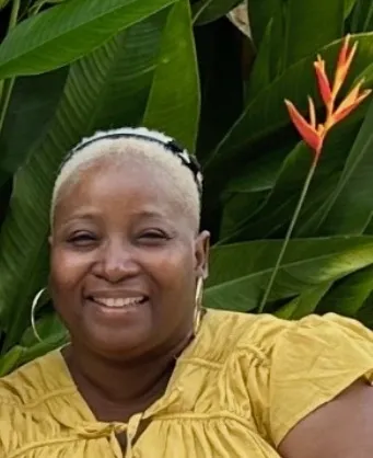 A smiling woman with short hair and hoop earrings poses in front of large green leaves.