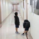 Two children wearing black clothes and facing away from the camera wait in a long hallway alone.