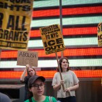 Protesters hold signs calling for the abolition of ICE and immigrant rights during a demonstration in front of a brightly lit display.
