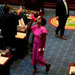 Florida state Rep. Angie Nixon, in a pink jumpsuit with a pink megaphone, stands on the floor of the Florida House amid people in suits at desks.