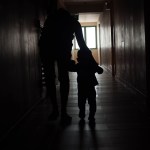 A silhouetted adult holds the hand of a young child as they walk down a dark hallway toward a window.