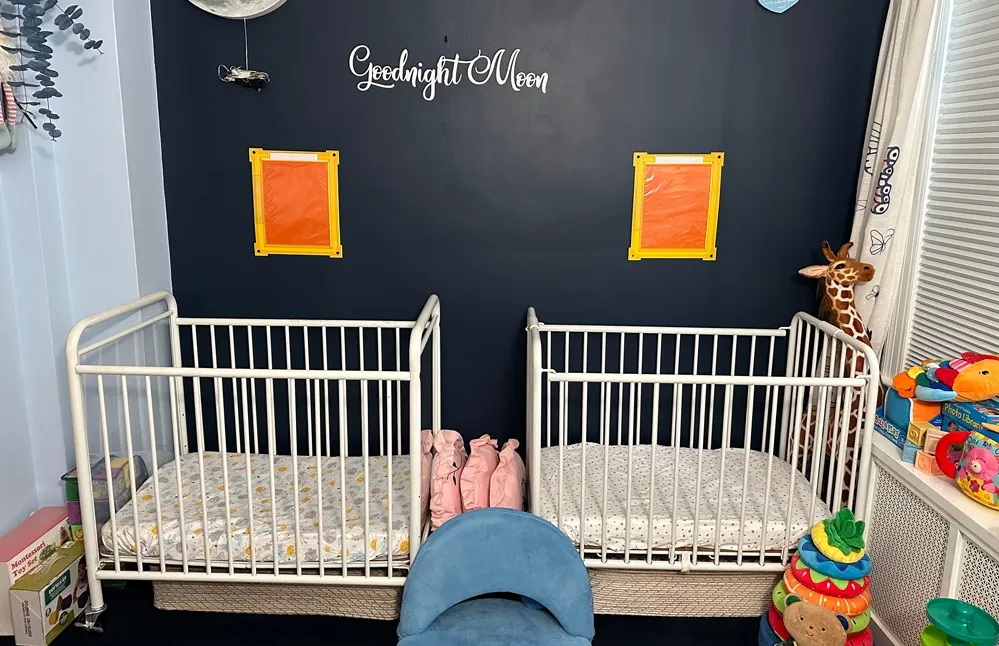 Two white cribs sit side by side in a home-based child care nursery, surrounded by children's toys and books beneath a "Goodnight Moon" wall decoration.
