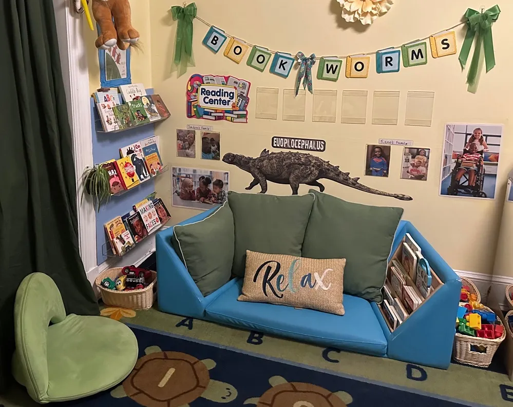 A home-based child care reading area features a blue couch, children's books, family photos and a dinosaur decoration beneath a "Book Worms" banner.