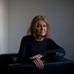 Gloria Steinem sits on a black chair and smiles softly as she poses for a portrait.