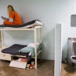 A woman in an orange uniform sits cross-legged on the top bunk of a metal bed reading a book. A stainless steel toilet and small mirror are mounted on the wall beside the bunk.